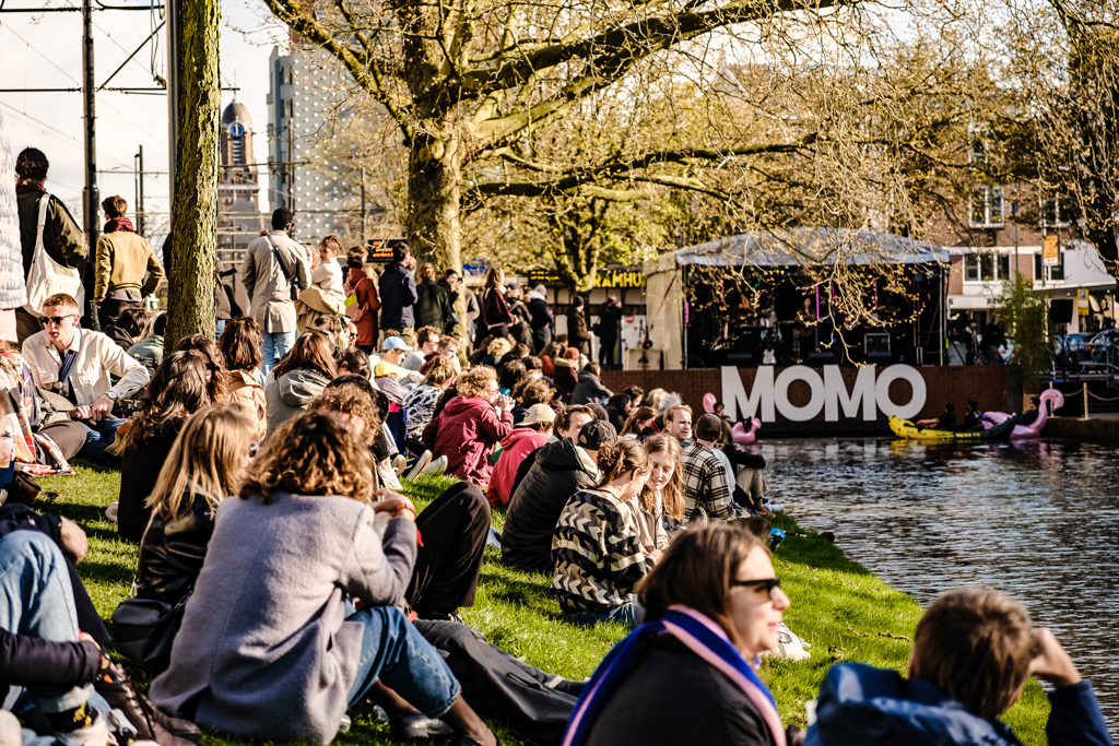 Publiek zit op het gras aan het water tijdens het MOMO Festival. Op de foto is water te zien, een podium en aan de linkerkant het gras waar het publiek op zit. 