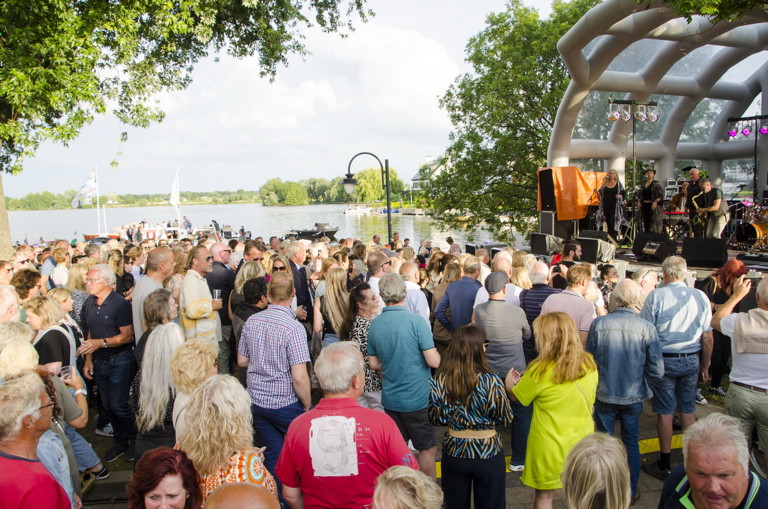 Foto van diverse mensen die voor een klein podium staan. Op het podium is een klein bandje aan het spelen. 