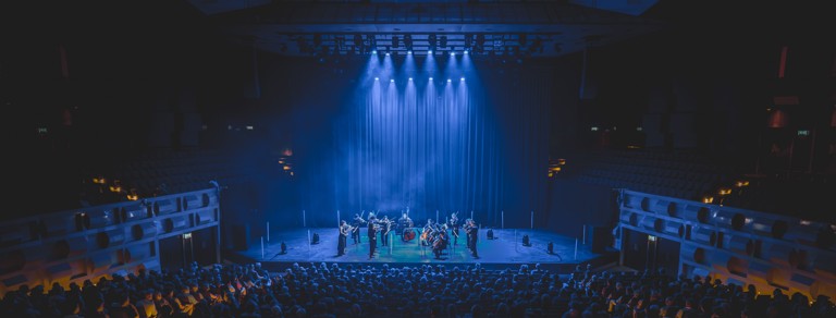 Foto van een optreden in de Doelen. Op het podium staat een klein orkest en de hele zaal zit vol met publiek. Er hangen lampen in de zaal die blauw licht afgeven waardoor er een hele blauwe gloed over de zaal heen gaat. 