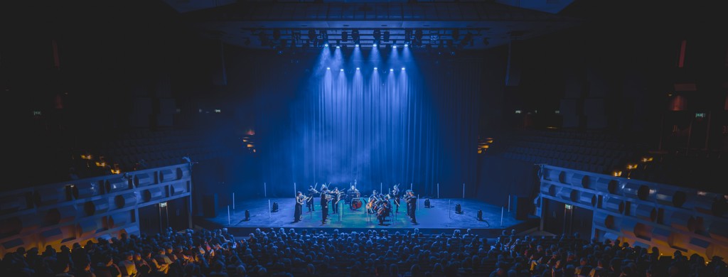 Foto van een optreden in de Doelen. Op het podium staat een klein orkest en de hele zaal zit vol met publiek. Er hangen lampen in de zaal die blauw licht afgeven waardoor er een hele blauwe gloed over de zaal heen gaat. 