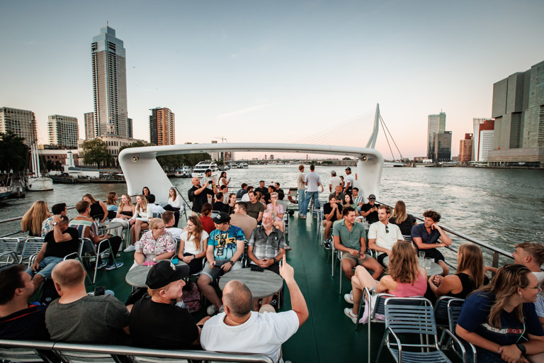 Foto van verschillende mensen op een boot. De boot vaart over water en op de achtergrond is de Erasmusbrug in Rotterdam te zien. 